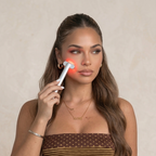 A woman modeling a brown and gold striped two-piece set, featuring a strapless crop top and matching high-waisted shorts with a subtle polka dot pattern. She is holding a white handheld device that emits a red light on her cheek, set against a neutral beige background.
